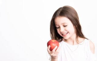 Positive young girl with brown hair in white clothes holding red apple and looking at it on white background